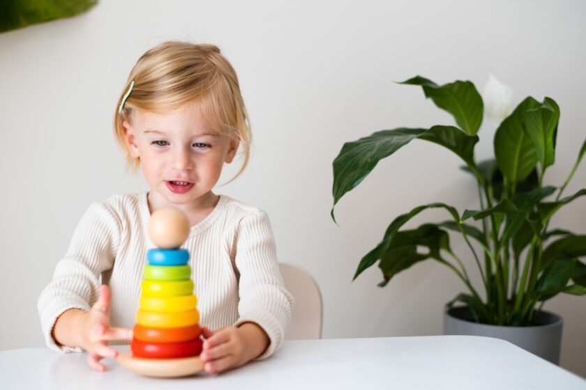 Young child sits at a table, playing with a colorful wooden stacking toy; a potted plant is visible in the background.