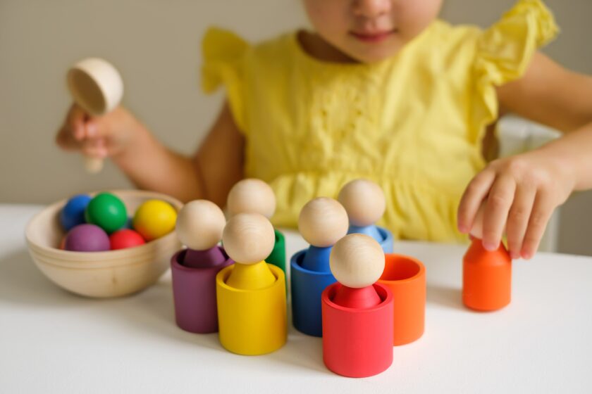 A child in a yellow dress plays with colorful wooden peg dolls and matching cups at a table.