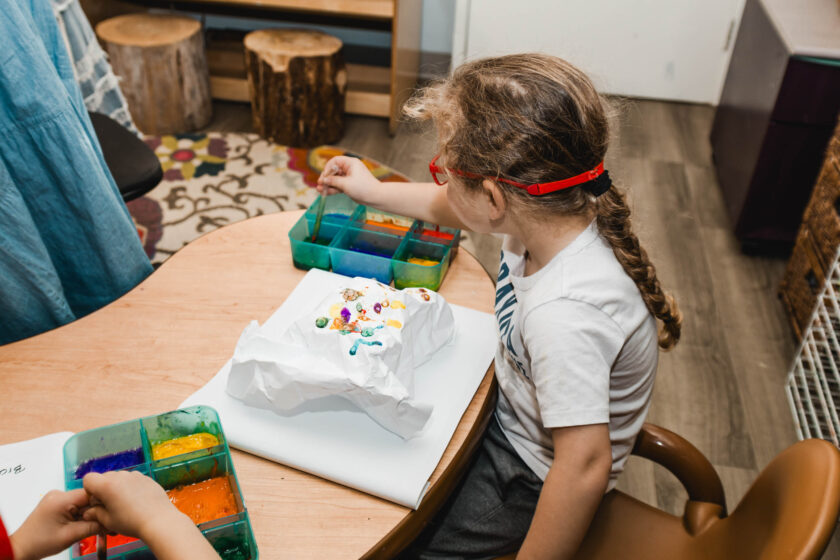 A child with braided hair and red glasses paints on a sheet of paper at a table using a tray with various paint colors.