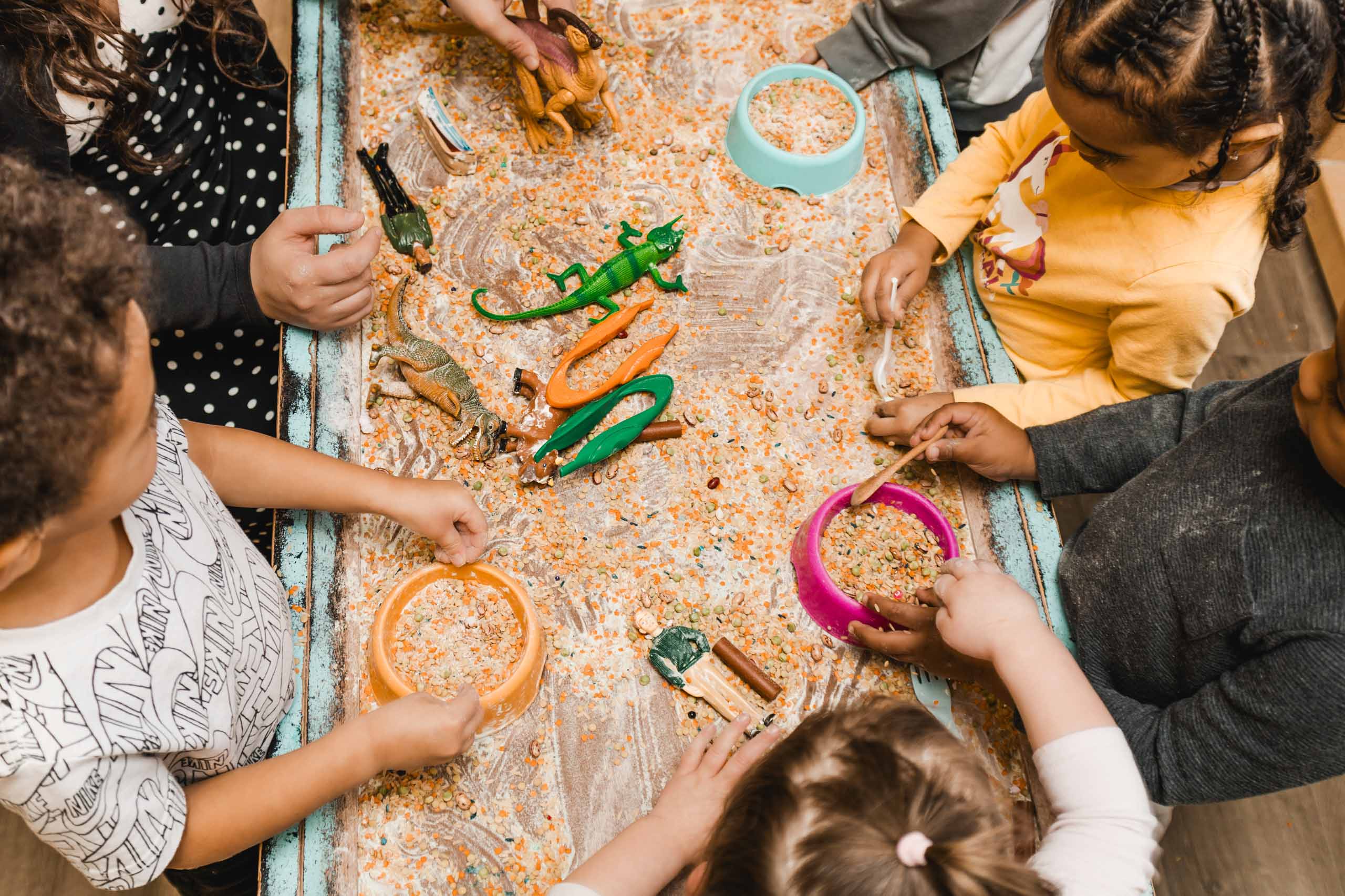 Four children play around a sensory table filled with rice, plastic dinosaurs, and bowls, using scoops and their hands to interact with the items.