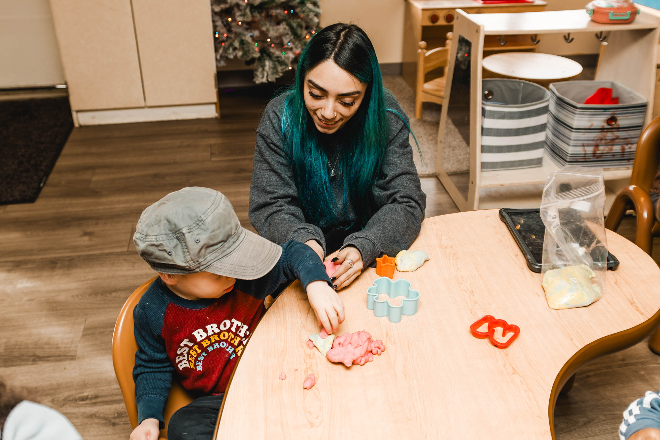 An adult and a child sit at a table playing with colorful playdough and plastic cutters in a classroom setting.