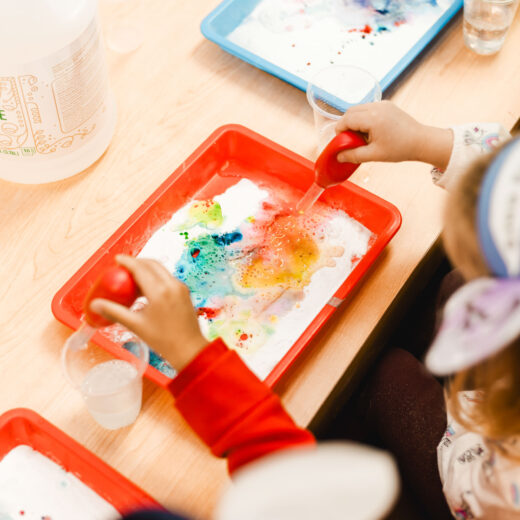 Children use droppers to mix colored liquids on trays of white substance, likely engaging in a science or art activity at a classroom table.