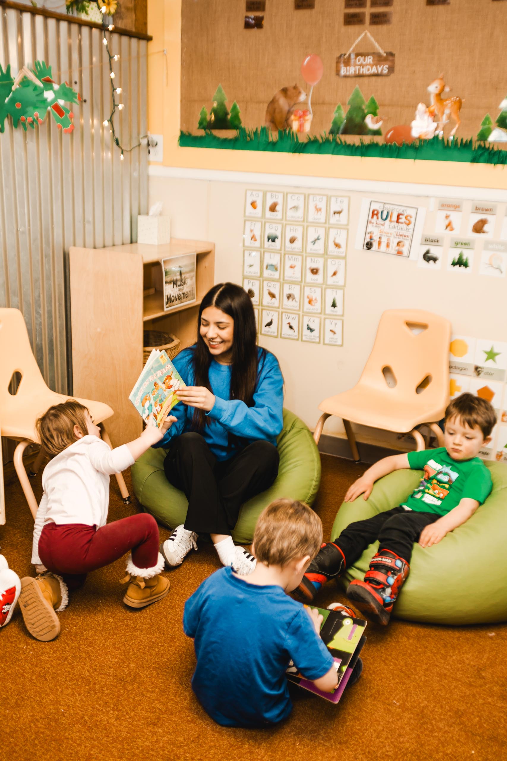 A woman reads a book to three young children in a classroom; two kids sit on bean bags, and one reaches toward the book. Educational posters decorate the walls.